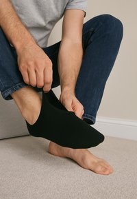 Sort ankelstrømpe bliver trukket på en fod, med synlig blød stofstruktur. Personens hånd griber strømpe, mens den anden fod er bar.