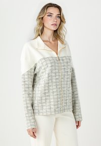 Veste zippée en crème avec un motif à carreaux texturé. Tissu doux avec des manches longues, un col et une fermeture éclair beige. Look minimaliste et moderne.