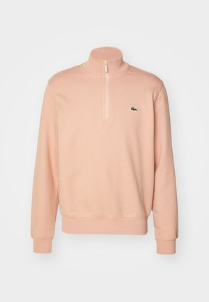 Lichtroze ritsvest met hoge kraag, ribbelmanchetten en -zoom. Bevat een klein groen logo op de borst. Zachte stoftextuur.