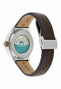 Reloj de acero inoxidable con correa de cuero marrón, fondo transparente, corona dorada y movimiento turquesa visible. Detalle de logotipo grabado presente.