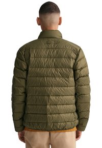 Chaqueta acolchada verde oliva con acolchado horizontal, cuello alto y un parche de logo en la parte posterior. Llevada sobre una camisa marrón.