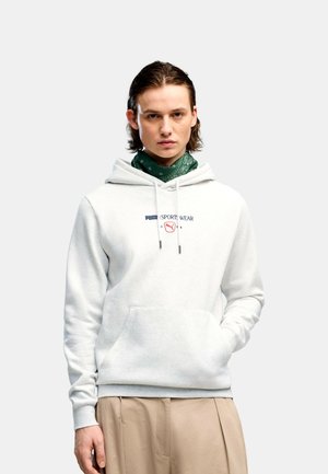 Junger Mann mit weißem Puma-Hoodie, grünem Halstuch und beigen Hosen, stehend vor einem einfarbigen hellgrauen Hintergrund.
