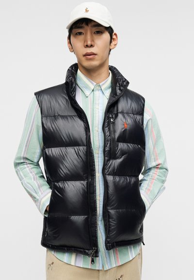 Fekete, magas nyakú, cipzáros előlapú és oldalzsebes pufi mellény, színes csíkos, gombos ing fölött viselve.