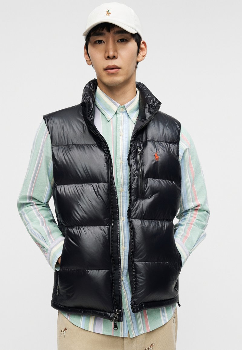 Veste matelassée noire avec un col montant, une fermeture éclair à l'avant et des poches latérales, portée sur une chemise à rayures multicolores boutonnée.