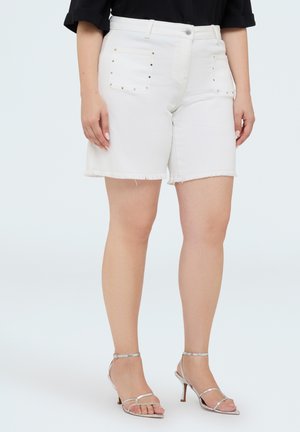 Vrouw draagt witte shorts met een hoge taille met gestudeerde voorzakken en zilveren sandalen met hoge hakken en bandjes, staand tegen een effen achtergrond.