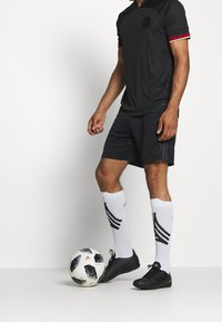 Uomo vestito con abbigliamento sportivo nero e calze bianche, in piedi con un piede su un pallone da calcio Adidas bianco e nero, su uno sfondo neutro.