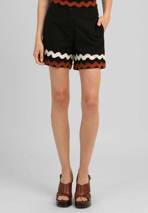 Femme portant un short noir avec un galon ondulé marron et blanc, des talons compensés ouverts marron, debout sur un fond uni.
