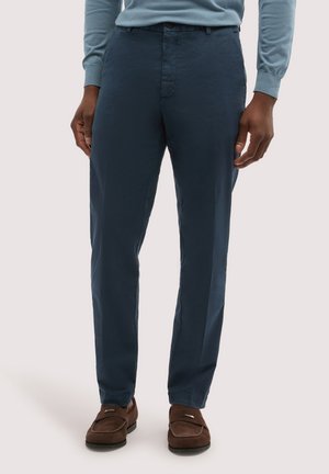 Mand iført navyblå bukser, lyseblå langærmet skjorte og brune ruskindssko, stående mod en ensfarvet baggrund.