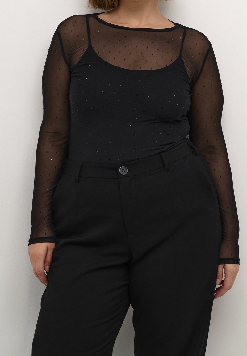 Femme portant un haut noir transparent à manches longues avec petits pois sur un caraco noir, associé à un pantalon noir taille haute.