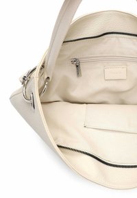 Kremowa skórzana torba wnętrze z zamykaną na suwak kieszenią, gładką fakturą i srebrnymi akcentami metalowymi. Otwarta, prezentująca przestronną podszewkę i przegródki.