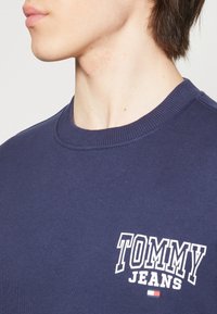 Felpa in cotone di un blu navy scuro con colletto a coste, presenta il logo "TOMMY JEANS" in bianco e un accento sotto il logo in rosso, bianco e blu.