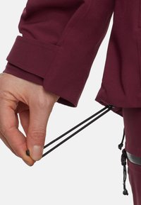 Mörk röd vattentät jacka med manschetter, med justerbara dragsnören vid fållen och en hand som greppar svarta snören.