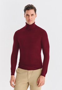 Burgundzki sweter z golfem, długi rękaw, wykonany z gładkiego, dopasowanego materiału; w połączeniu z jasnobeżowymi spodniami.