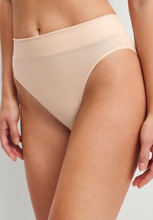 Vrouw die beige katoenen lingerie met hoge taille draagt, staat met ontspannen handen langs haar zij tegen een effen achtergrond.