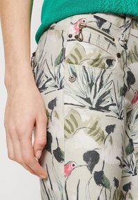 Main tenant posée à côté d'un pantalon à motifs avec des designs d'oiseaux et de feuilles, montrant une poche zippée et des clous en métal sur la ceinture.