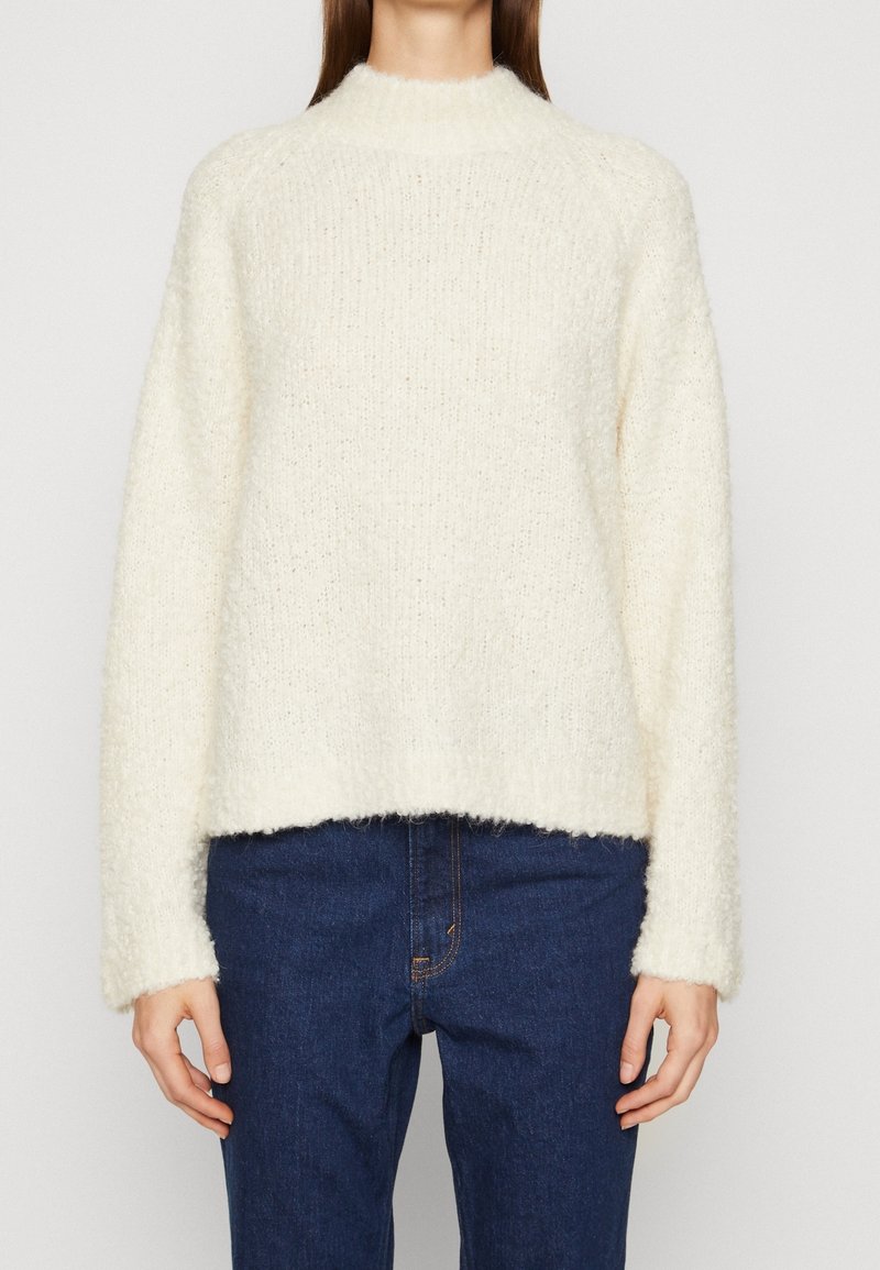 Kermanvärinen, teksturoitu neulepusero korkealla kauluksella ja laskostetuilla harteilla; väljä istuvuus, lyhyt pituus. Yhdistetty tummansinisten farkkujen kanssa.