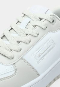 Ανοιχτό γκρι αθλητικό παπούτσι από συνθετικό δέρμα, με επίπεδα μπεζ κορδόνια, λευκές λεπτομέρειες, υφασμένη σόλα και έντονη λεπτομέρεια λογότυπου.