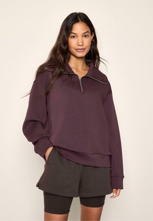 Giovane donna con lunghi capelli scuri che indossa una felpa con zip viola scuro e pantaloncini neri a strati, in piedi su uno sfondo neutro.