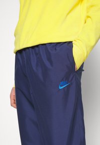 Granatowe spodnie sportowe z elastycznym paskiem w talii, z niebieskim logo Nike na lewej udzie. Materiał wygląda na gładki i lekki.