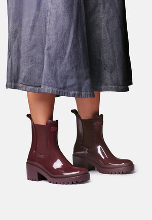 Bottines brillantes rouge foncé à talons épais portées avec une jupe en jean courte, dévoilant les mollets sur un fond uni clair.