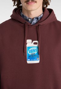 Röd hoodie med en grafisk bild av en blå och vit flaska märkt "VANS Liquid Off the Wall" tryckt på bröstet. Tyget verkar slätt.