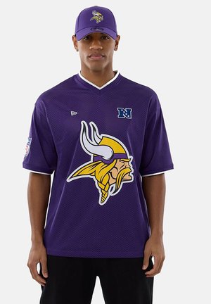 Man wearing a purple Minnesota Vikings cap and jersey with large Viking logo, standing against a plain white background.