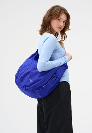 Junge Frau mit hellbraunen Haaren trägt ein hellblaues Langarmshirt und schwarze Hosen, sie trägt eine große, leuchtend blaue Umhängetasche.