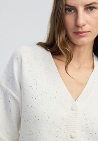 Młoda kobieta ubrana w białą dzianinową kardigan z małymi, rozproszonymi srebrnymi cekinami i guzikami pokrytymi tkaniną, z jasnobrązowymi włosami opadającymi na ramię.