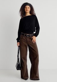 Γυναίκα με σγουρά μαλλιά που φοράει μαύρο τοπ με μακριά μανίκια, καφέ παντελόνι με φαρδιά πόδια, μαύρη ζώνη, κορδέλα και κρατάει μαύρη τσάντα χειρός.