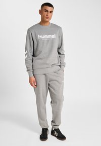 Mężczyzna ubrany w dopasowany szary dres hummel, czarne trampki, stoi z jedną ręką w kieszeni na jednolitym tle.