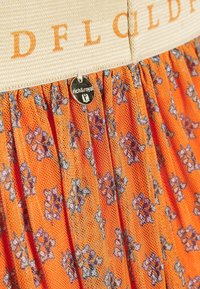 Orange plisserad tyg med små blå blommönster, beige midjeband med orange text, och en liten rund metalltag "rich&royal".