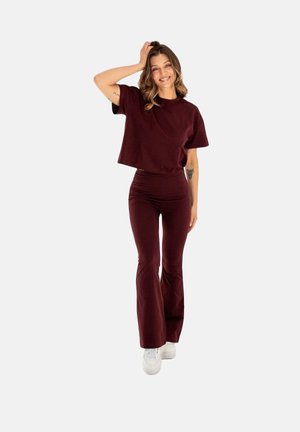 Młoda kobieta uśmiechająca się, ubrana w burgundową bluzkę z krótkim rękawem i dopasowane, rozkloszowane spodnie oraz białe tenisówki, stojąca na tle jednolitego tła.