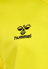 Jasnożółta koszula z poliestru z czarnym logo Hummel z wzorem pszczoły. Tekstura jest gładka, a fason sportowy.