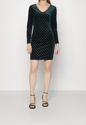 Femme vêtue d’une robe en velours vert foncé ajustée à manches longues avec des motifs en grille argentés et des sandales noires à talons hauts, debout devant un fond uni.