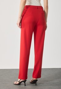 Rode wijde broek van soepele stof met een tailleband, met een achterzak. Gepaard met zwarte sandalen met bandjes op een grijze vloer.