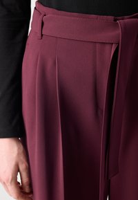 Burgunderfargede bukser med høy midje og legg, et matchende belte og en glatt tekstur. Stylet med en svart langermet topp.