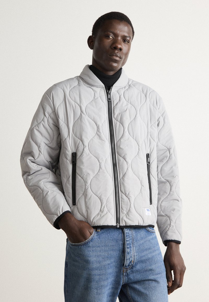 Light gray quilted bomber jacket with a stand collar, zip front, and black zipper accents. Features a textured, padded surface and elastic cuffs.