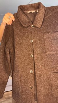 Hand håller i en brun knäppt jacka med krage och två framfickor, visas inomhus mot en bakgrund av ljust trägolv.