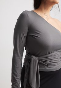 Haut asymétrique gris à manches longues avec un lien de taille froncé, fabriqué en tissu doux et extensible. Texture lisse, design minimaliste.