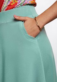 Tyrkysová sukně s texturovanou látkou a boční kapsou. Ruka je vidět opřená v kapse, na které je zlatý náramek z řetízků.