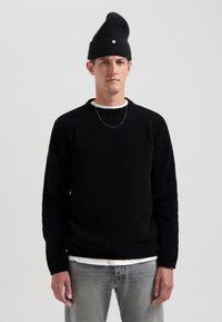 Schwarz strukturiertes Strick-Sweatshirt mit Raglanärmeln und Rundhalsausschnitt, kombiniert mit einem weißen Shirt, Lichtgraue Jeans und einer schwarzen Mütze.