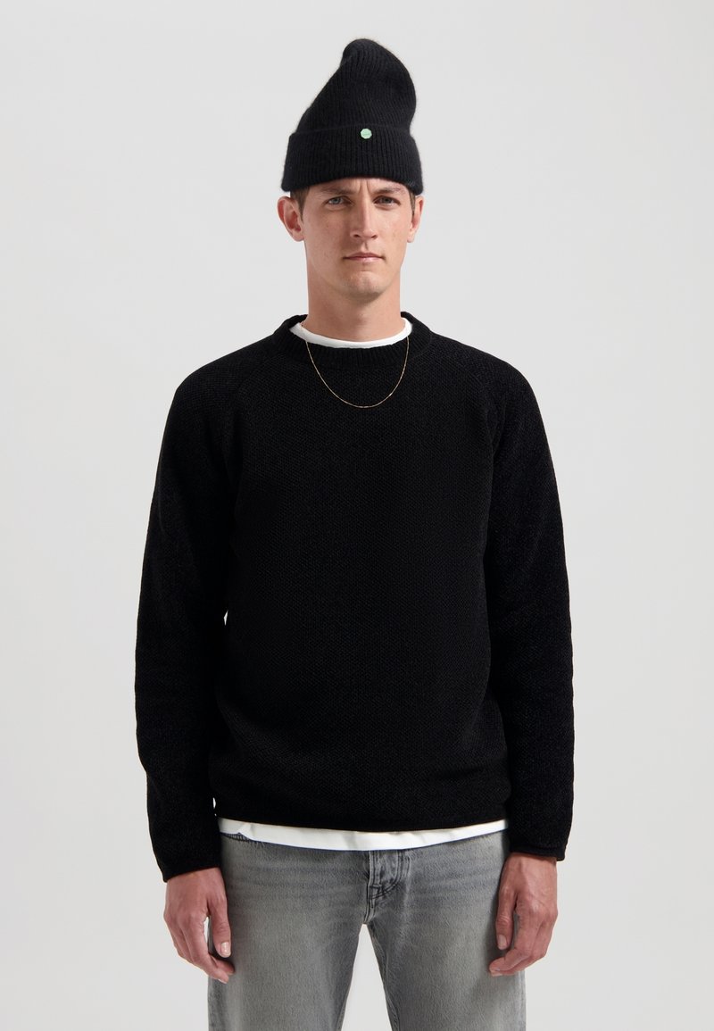 Schwarz strukturiertes Strick-Sweatshirt mit Raglanärmeln und Rundhalsausschnitt, kombiniert mit einem weißen Shirt, Lichtgraue Jeans und einer schwarzen Mütze.