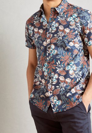 Mężczyzna ubrany w granatową, kwiecistą koszulę z krótkim rękawem z niebieskimi, białymi i rdzawego koloru kwiatami, ręce w kieszeniach ciemnych spodni.