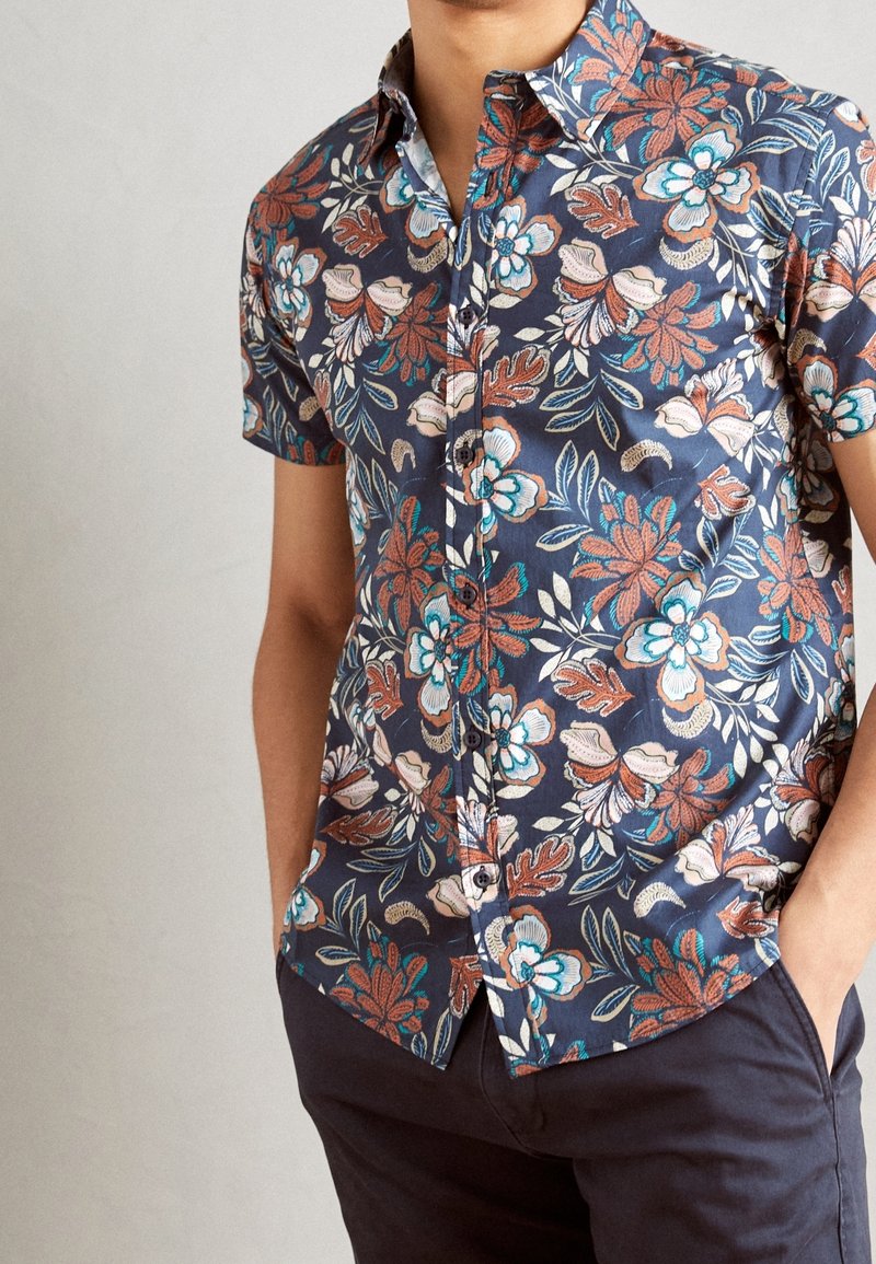Homme portant une chemise boutonnée à manches courtes bleu marine à fleurs bleues, blanches et couleur rouille, mains dans les poches d’un pantalon sombre.