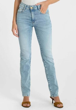 Femme portant un jean slim taille haute bleu clair et des sandales à talons ouvertes marron, debout avec une main dans la poche.