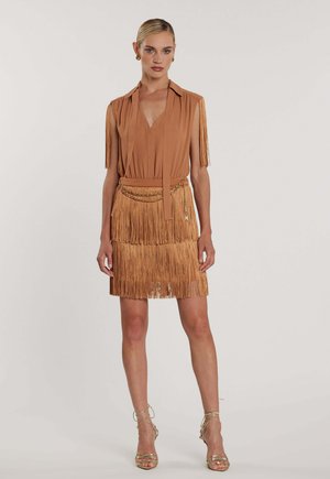 Donna che indossa un vestito color sabbia con frange e gonna a strati, design senza maniche con spalle scoperte, cintura a catena dorata e tacchi dorati con cinturini, in piedi su sfondo semplice.