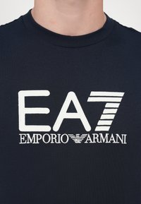 T-shirt in cotone blu navy con testo ricamato bianco "EA7 EMPORIO ARMANI," con dettaglio a righe nel numero "7." Semplice scollatura a girocollo.