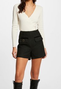Femme portant un pull crème à col en V côtelé, rentré dans un short noir texturé taille haute avec des poches à rabat boutonnées, et des bottes noires montant jusqu'aux genoux.