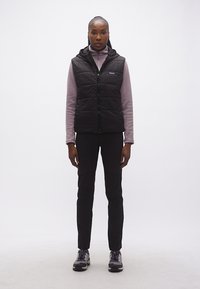 Γυναίκα όρθια φοράει μαύρο μπουφάν χωρίς μανίκια πάνω από μωβ μπλουζάκι με μακριά μανίκια, μαύρο παντελόνι και γκρι παπούτσια πεζοπορίας σε λευκό φόντο.