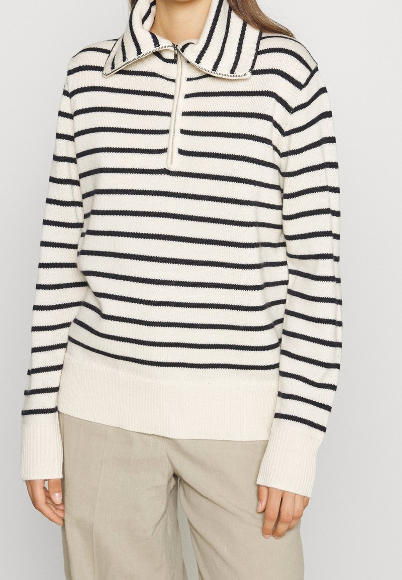 Femme portant un pull en tricot blanc avec des rayures horizontales noires, un col à demi-fermeture éclair, et un pantalon beige, debout devant un fond uni.